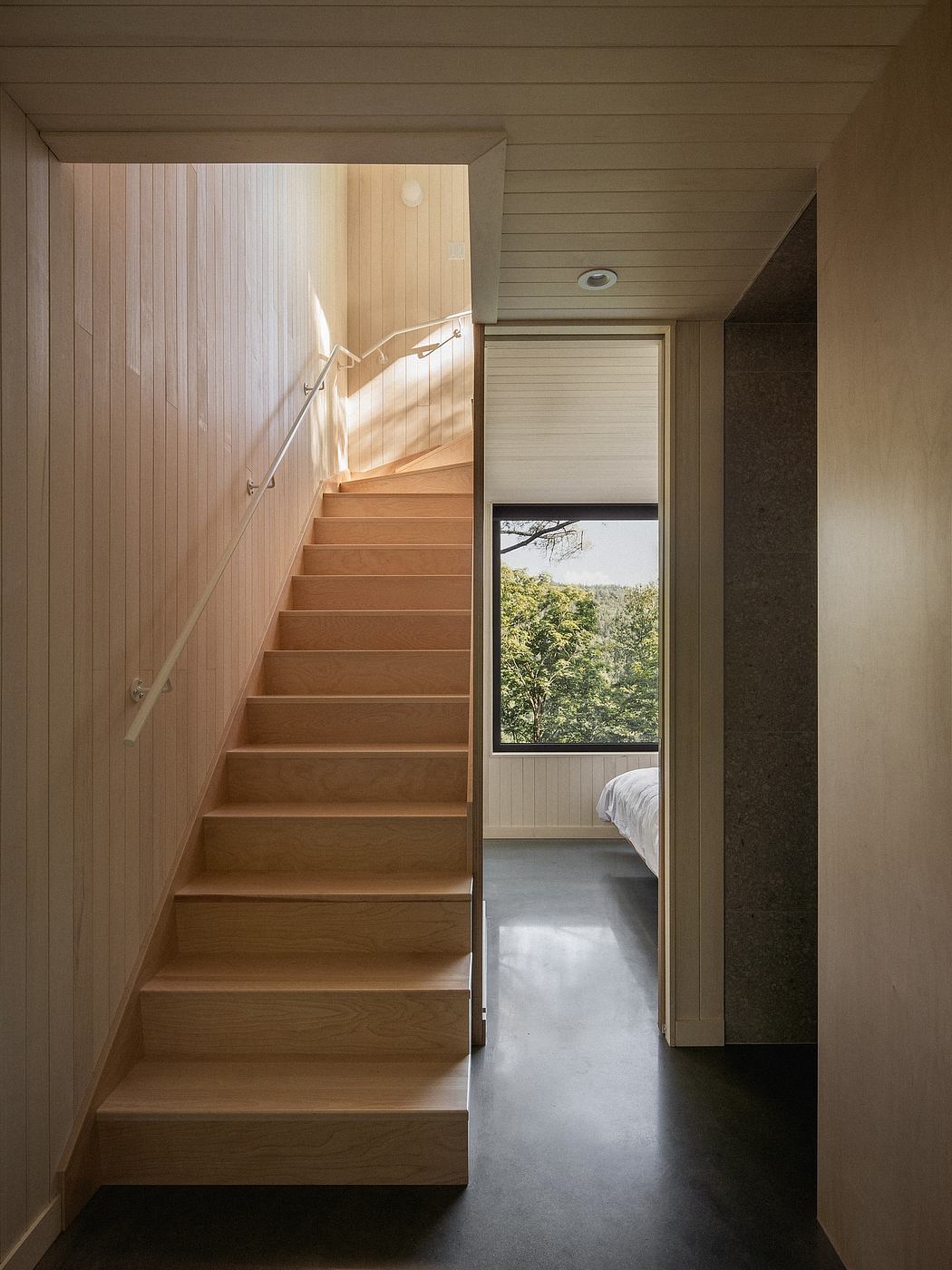 Wooden stairs and paneling lead to a bedroom with a view of the lush greenery outside.