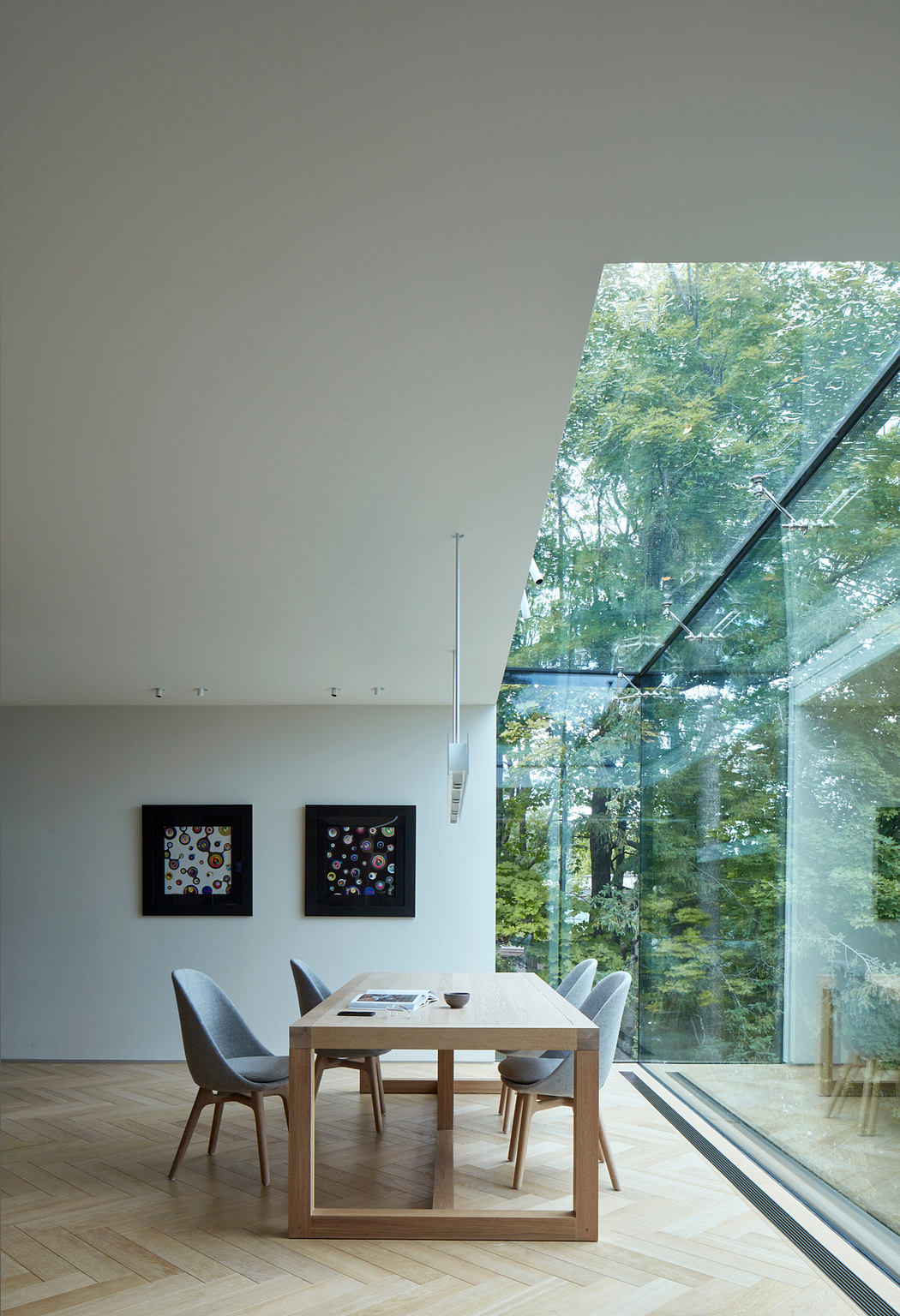 A bright, modern dining room with a wooden table, minimalist chairs, and a panoramic glass wall overlooking a lush forest.