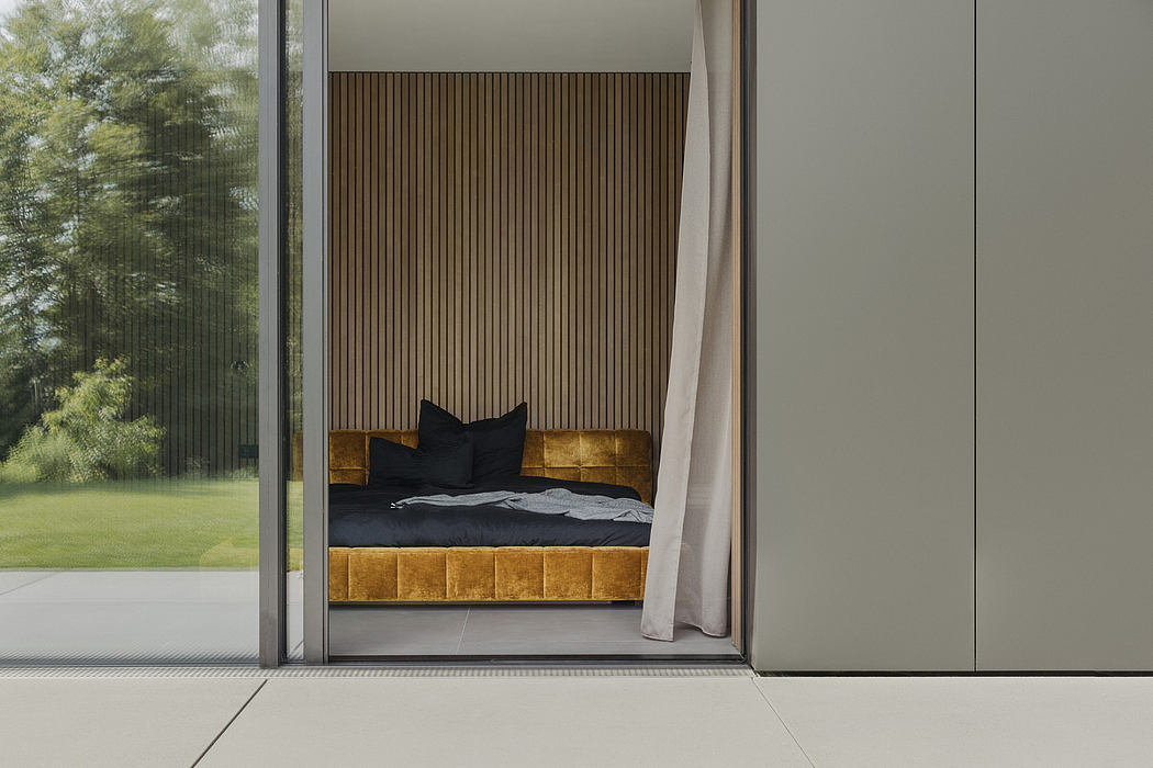 Modern bedroom with wooden wall paneling, gold upholstered bed, and floor-to-ceiling windows overlooking lush greenery outside.