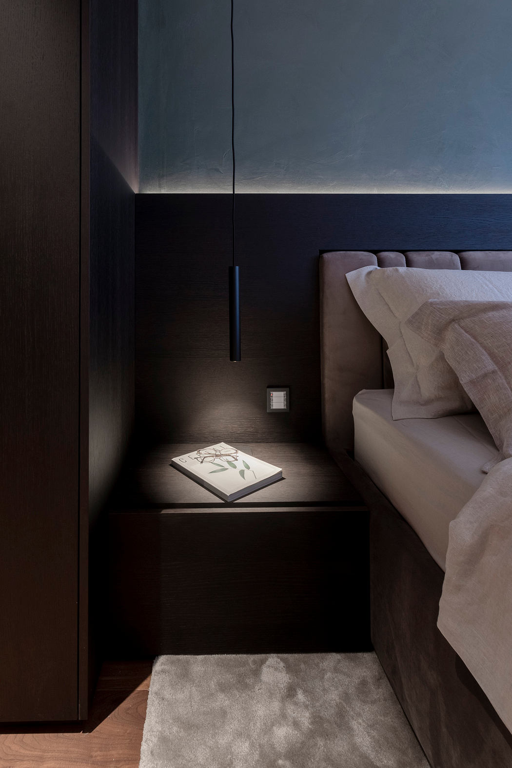 A dark, minimalist bedroom with a book on the nightstand and a sleek, modern lighting fixture.