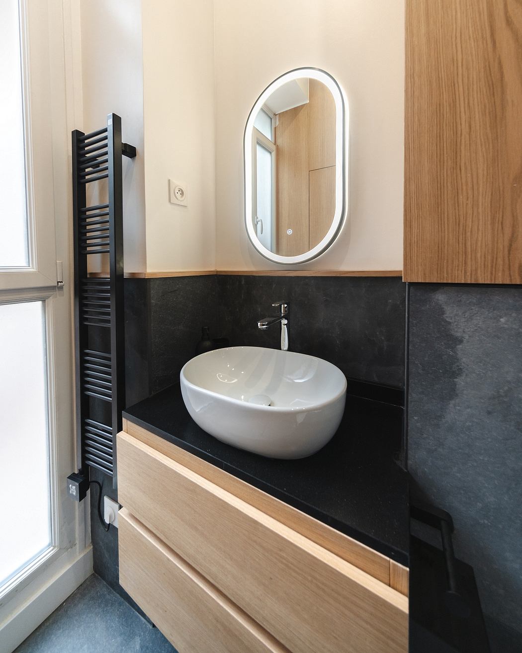 A modern bathroom with a sleek oval mirror, wood vanity, and a black stone countertop.