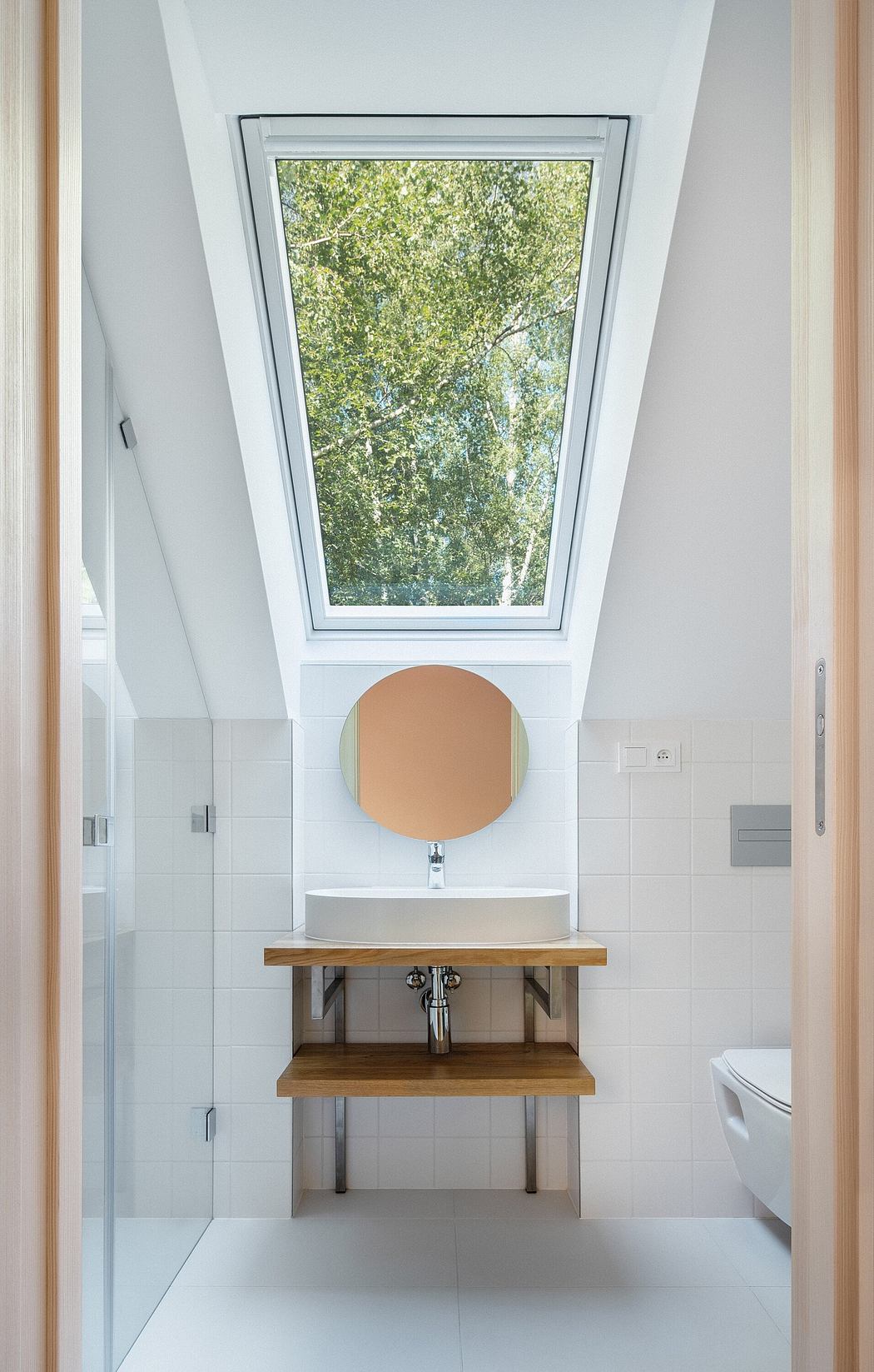 A modern bathroom with a circular mirror, wooden vanity, and large skylight window overlooking trees.