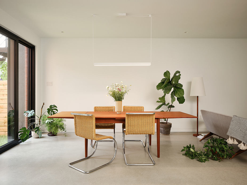 A bright, minimalist dining room with a wooden table, wicker chairs, and lush potted plants.