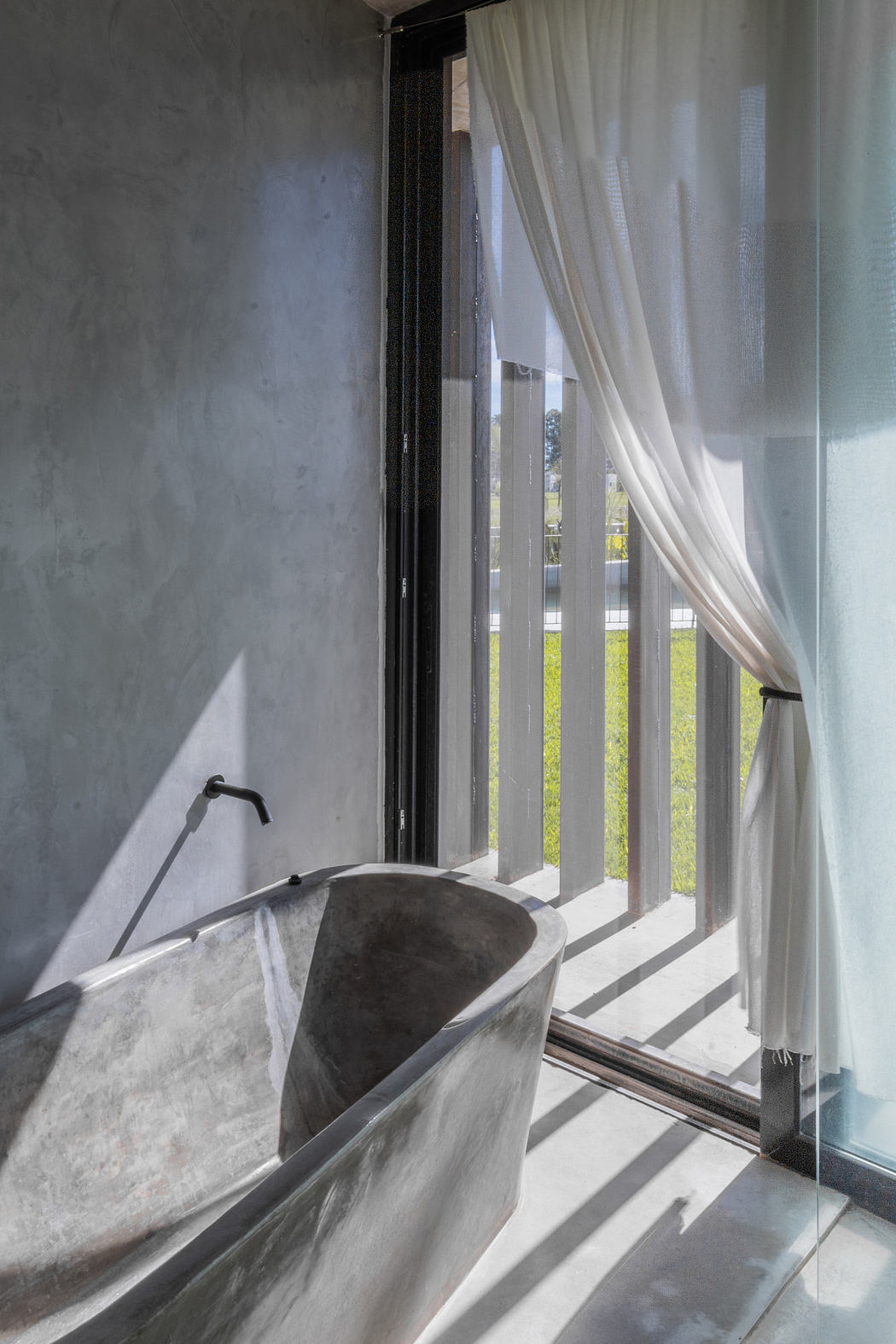 A minimalist bathroom with a concrete bathtub, floor-to-ceiling windows, and sheer curtains.