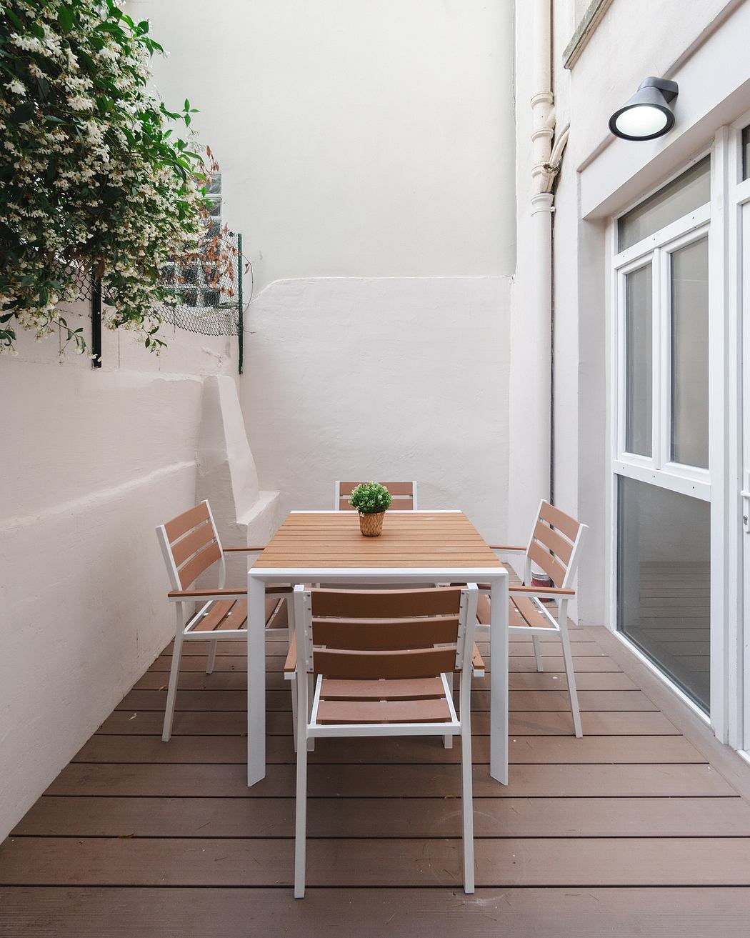 Small outdoor dining area with wooden table and chairs, white walls, and lush greenery.