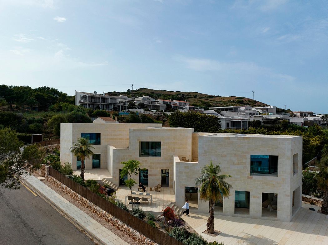 Modern beige stone building with large windows and palm trees, surrounded by lush landscape.