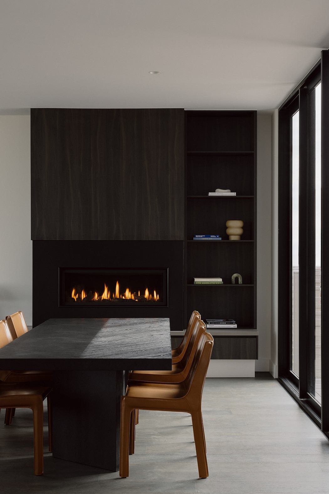 A minimalist dining room with a sleek black fireplace, built-in shelving, and large windows.