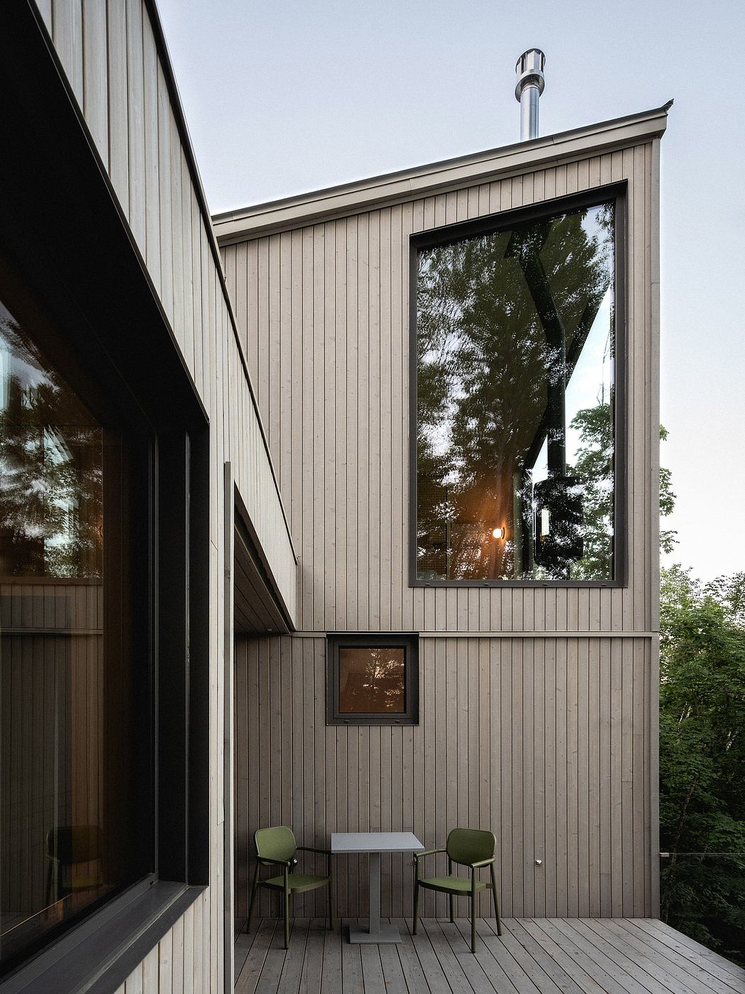 Modern wooden structure with large glass window, small deck, and green chairs.