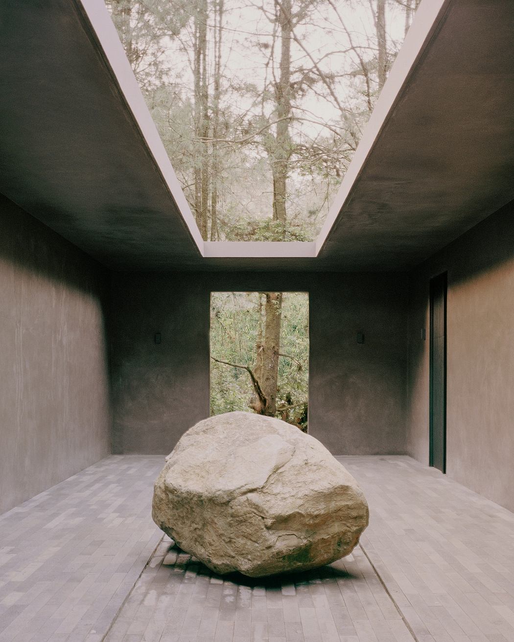 A large stone sculpture sits on a wood floor, surrounded by windows framing a forested view.
