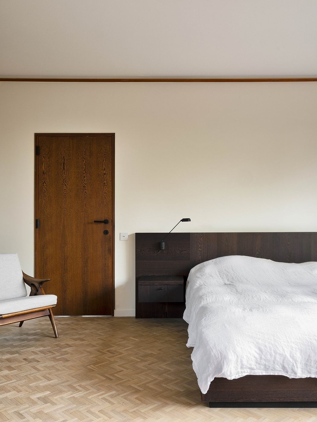 Minimalist bedroom with dark wooden features, white bedding, and a mid-century inspired chair.