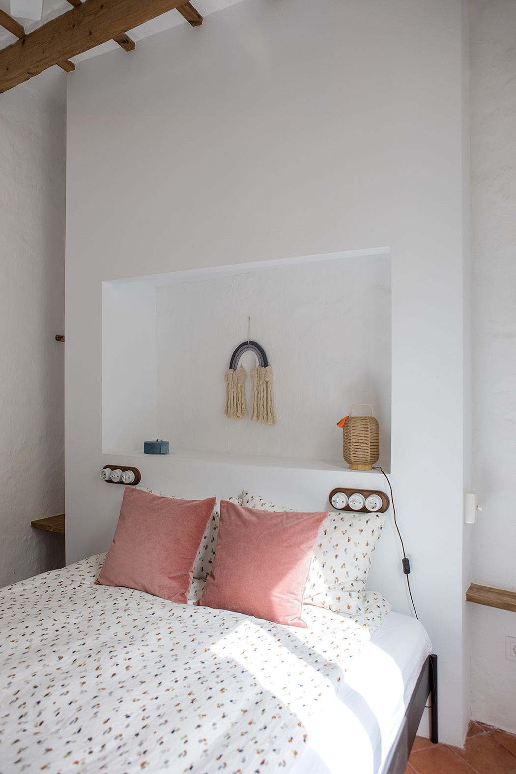 Cozy bedroom with exposed wooden beams, white walls, and decorative macrame wall hanging.
