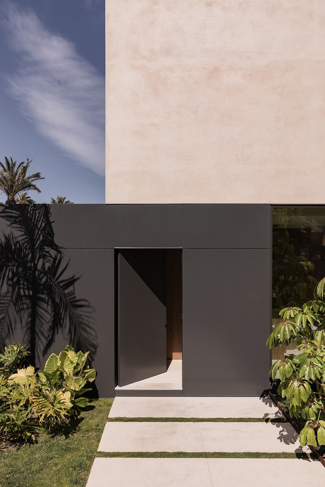 The image shows a modern, minimalist building with a black exterior and a prominent entryway surrounded by lush tropical foliage.