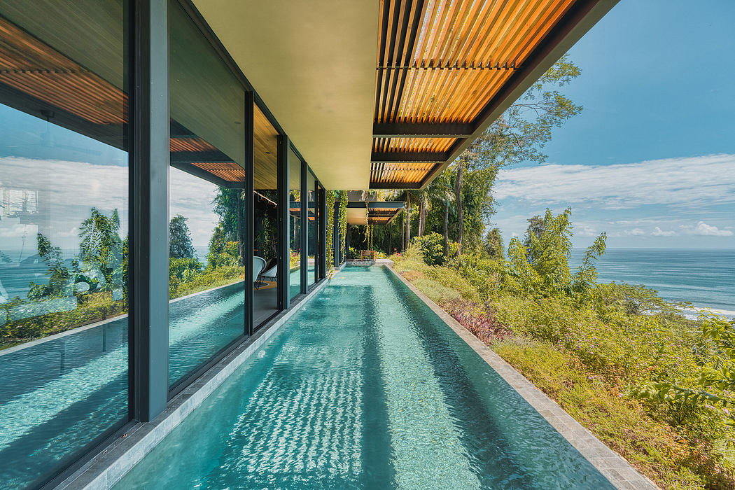 A modern, glass-enclosed hallway with a long pool running along its length, surrounded by lush greenery.