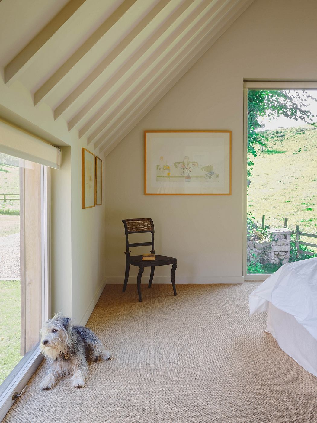 Bright, airy bedroom with wooden beams, framed artwork, and cozy chair overlooking nature.