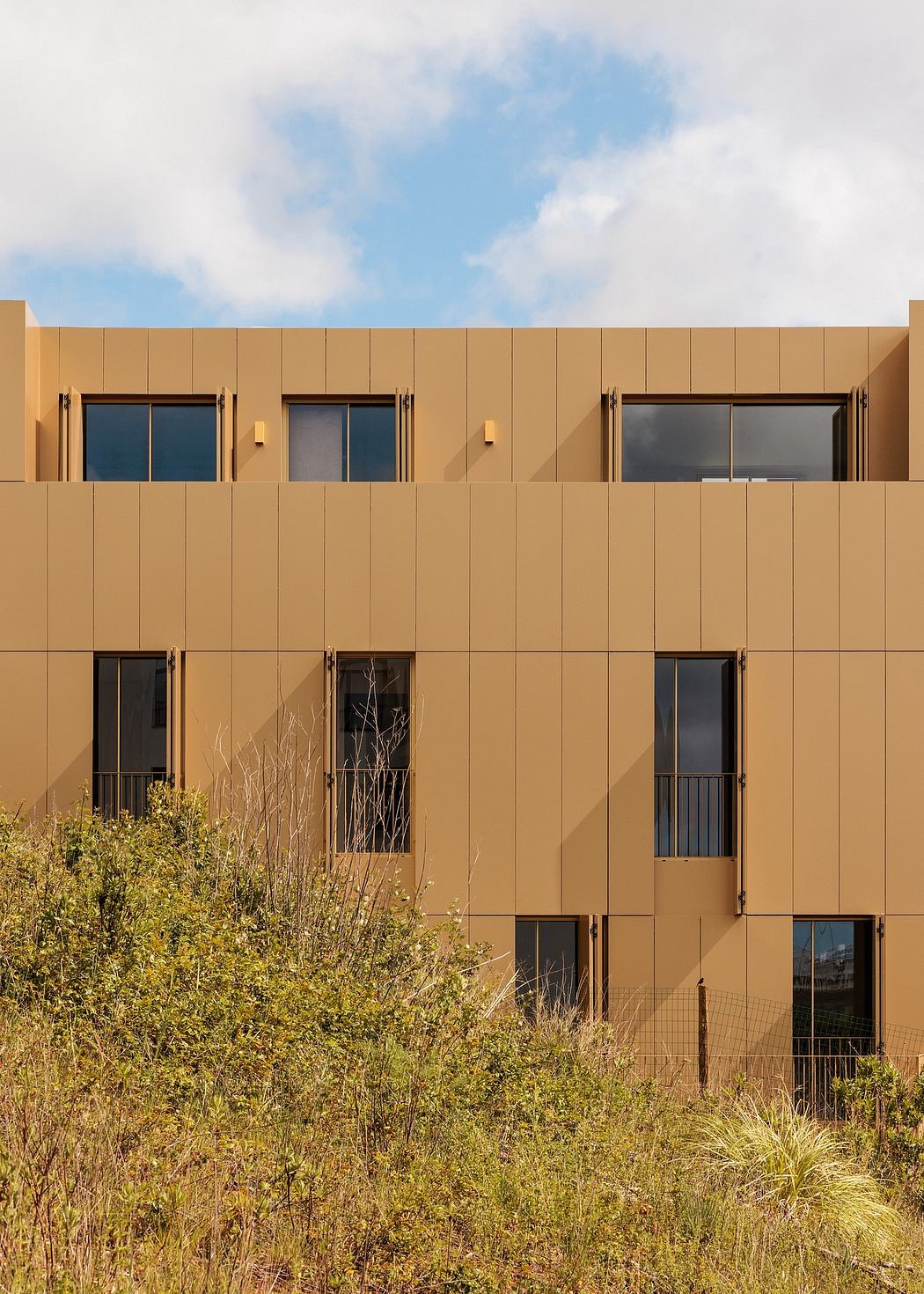 A modern, ochre-colored building with uniform windows and balconies, surrounded by lush vegetation.