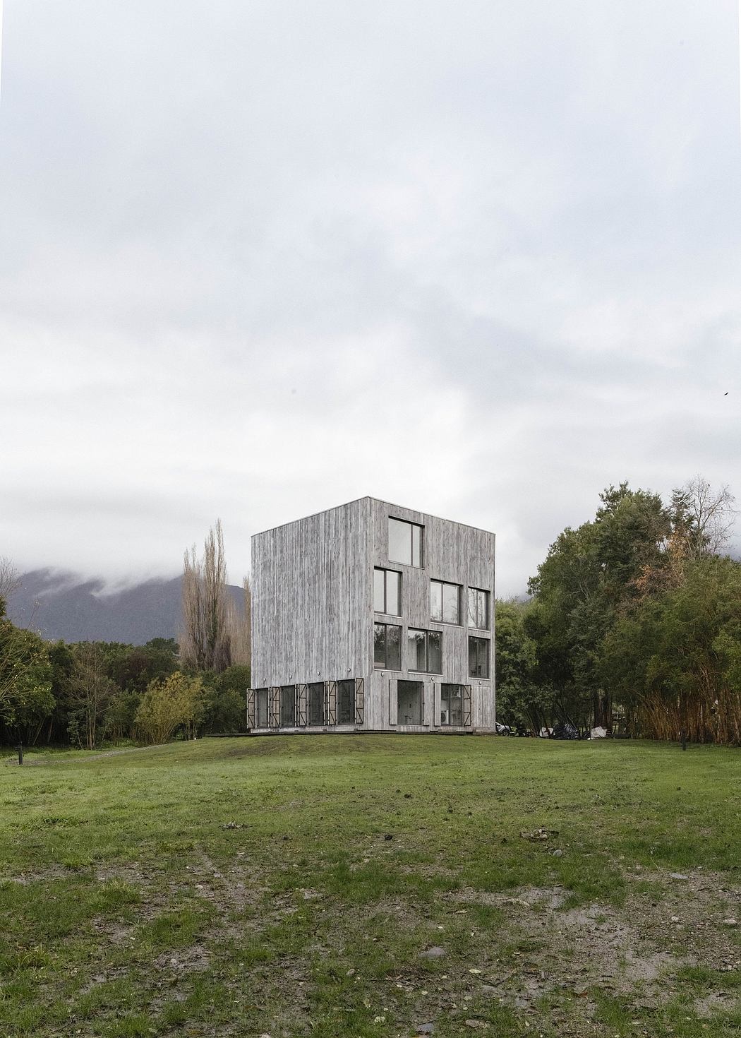 A modern, rectangular wooden structure with large windows stands in a grassy field surrounded by trees.