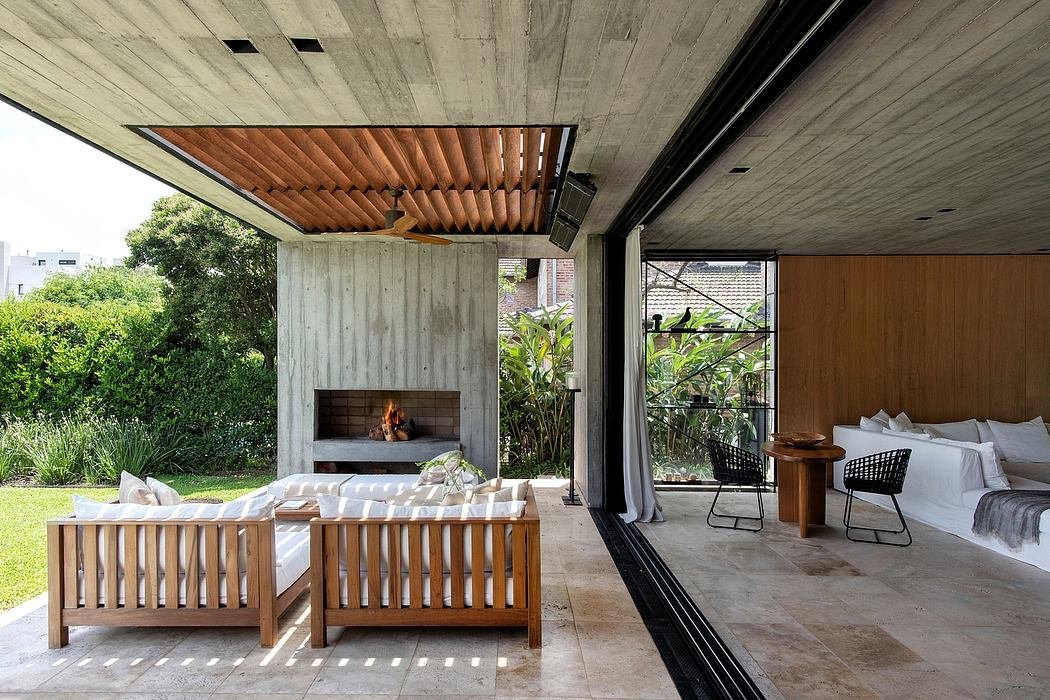Architectural outdoor lounge with concrete ceiling, wooden beams, fireplace, and lush vegetation.