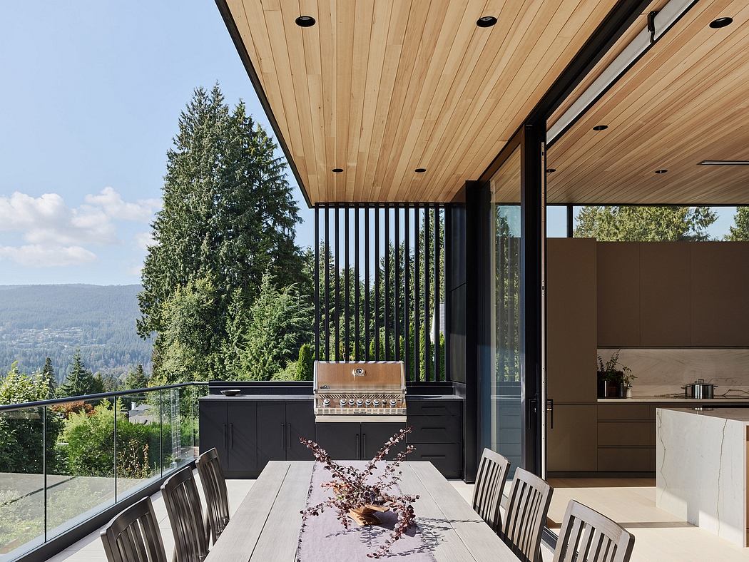 A modern outdoor kitchen with a wood-paneled ceiling, glass railings, and a dining area overlooking a forested landscape.