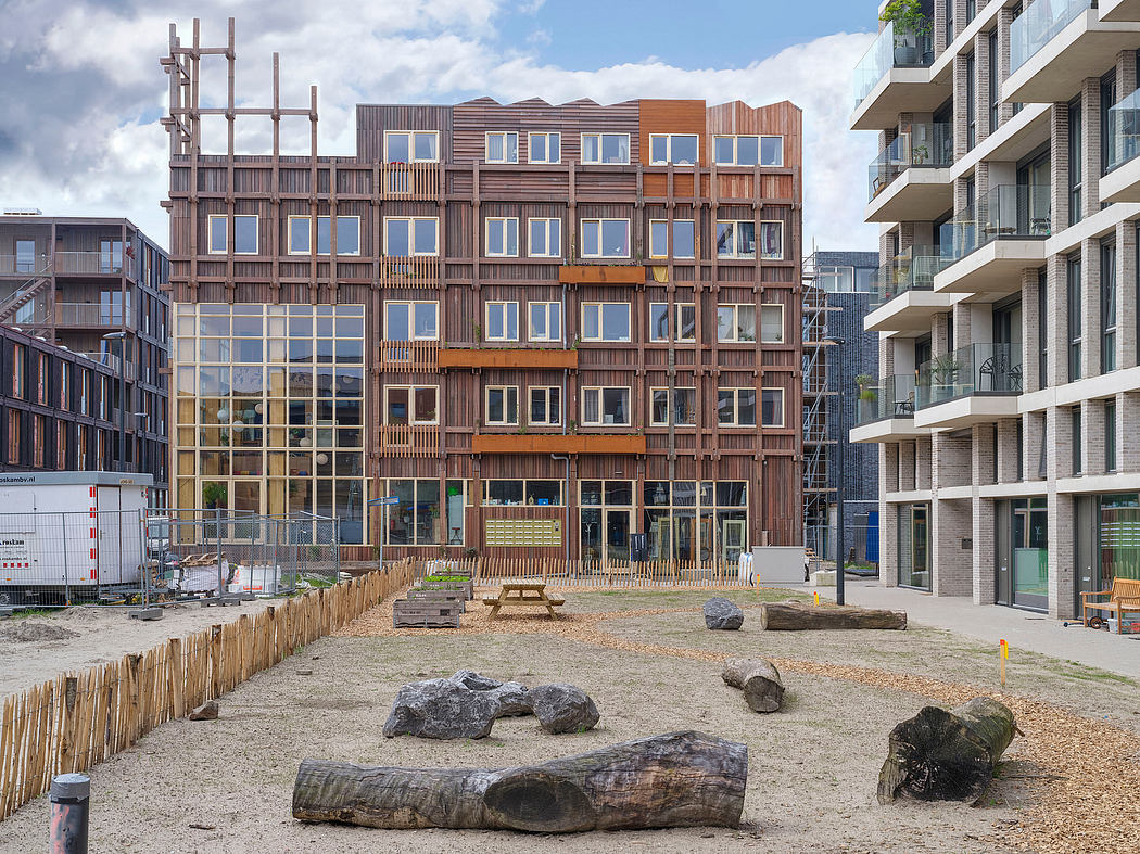 A modern multi-story building with a wooden facade and varied window patterns, surrounded by a gravel courtyard with natural elements.