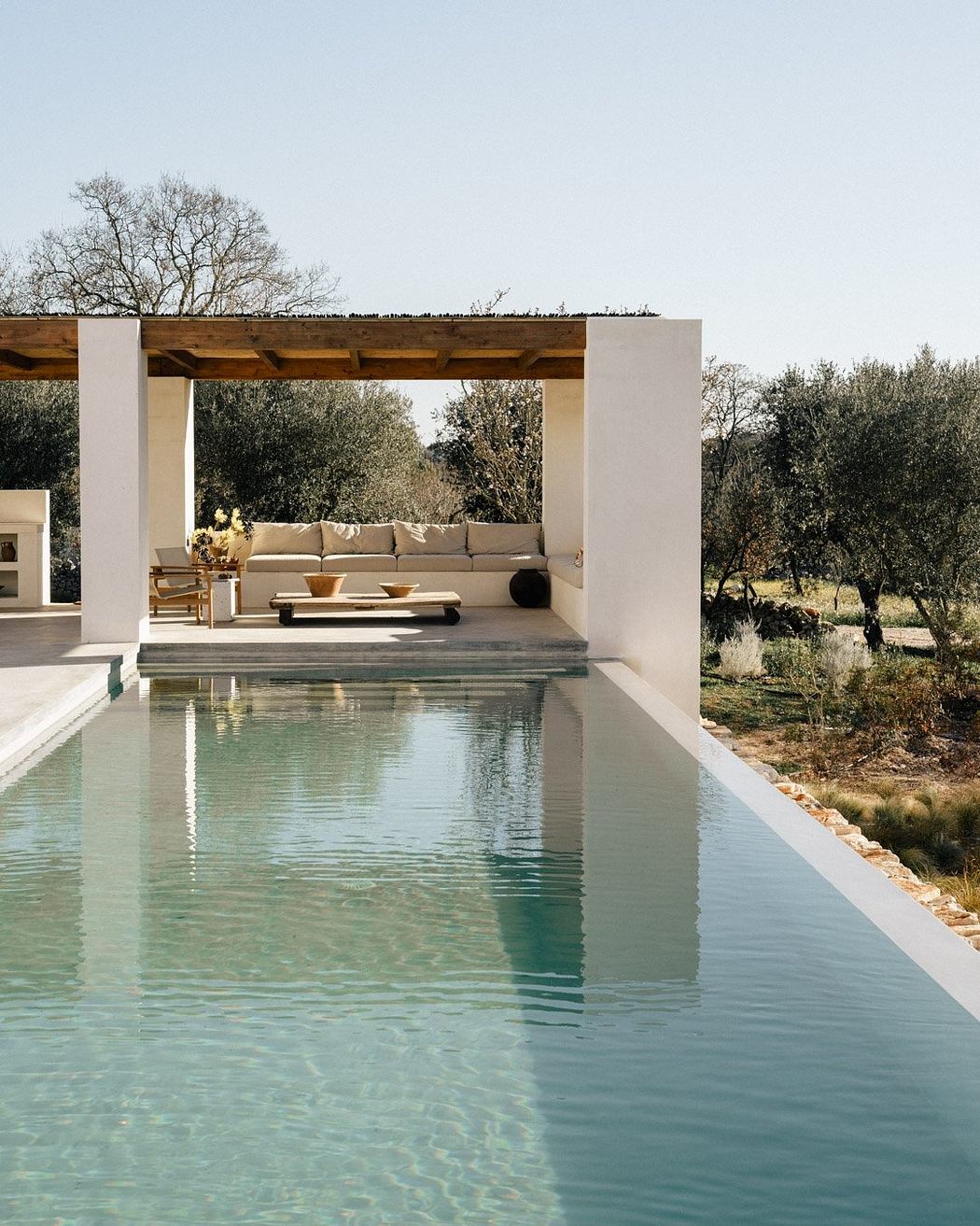 Minimalist outdoor patio with pool, wooden beams, and lush greenery backdrop.