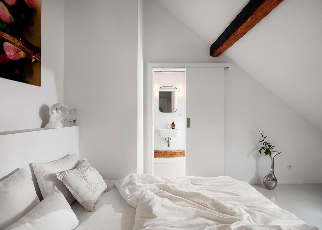 Cozy attic bedroom with white walls, wooden beams, and a partially visible bathroom.