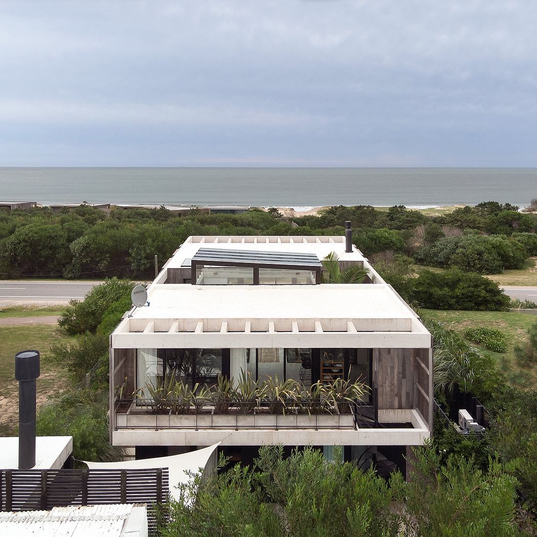 Modern beach house with large balcony, glass walls, and lush greenery surrounding it.