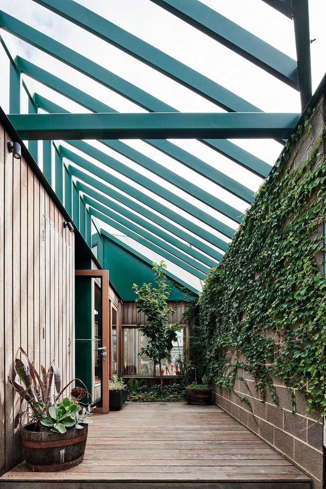 Wooden deck with potted plants, surrounded by a green-tiled roof and brick walls covered in vines.