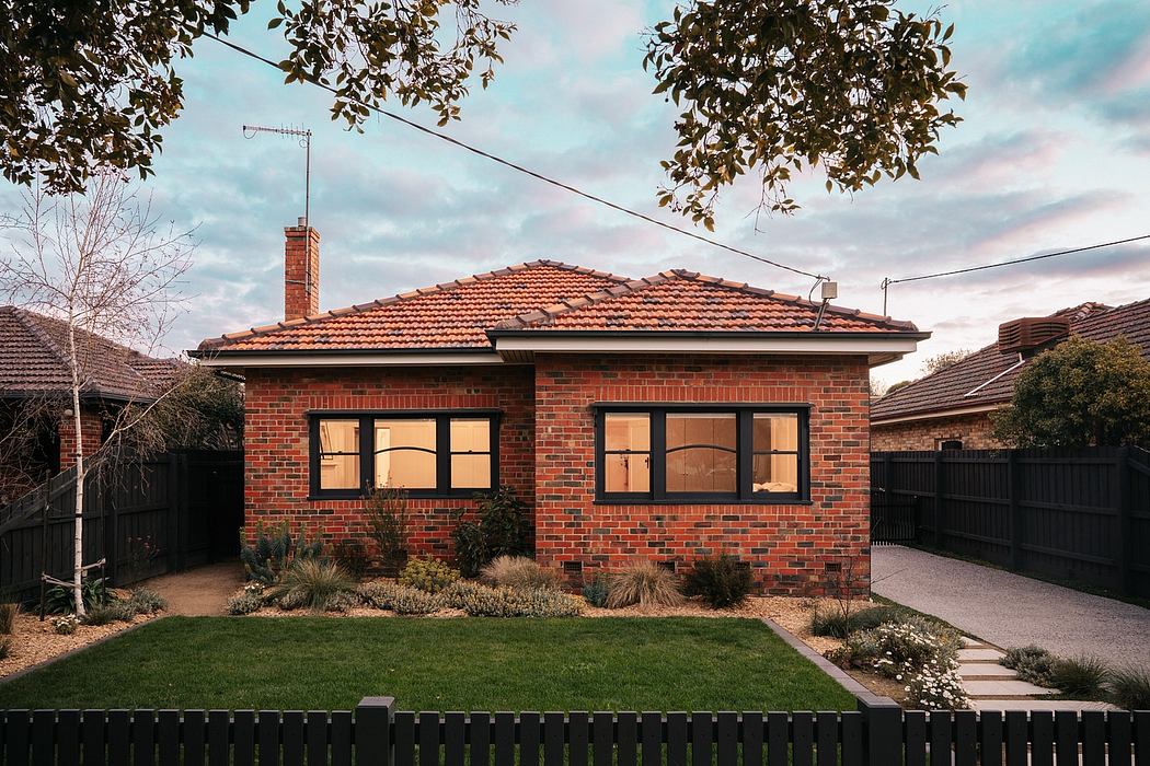 A cozy brick house with a tiled roof, surrounded by lush greenery and a manicured lawn.