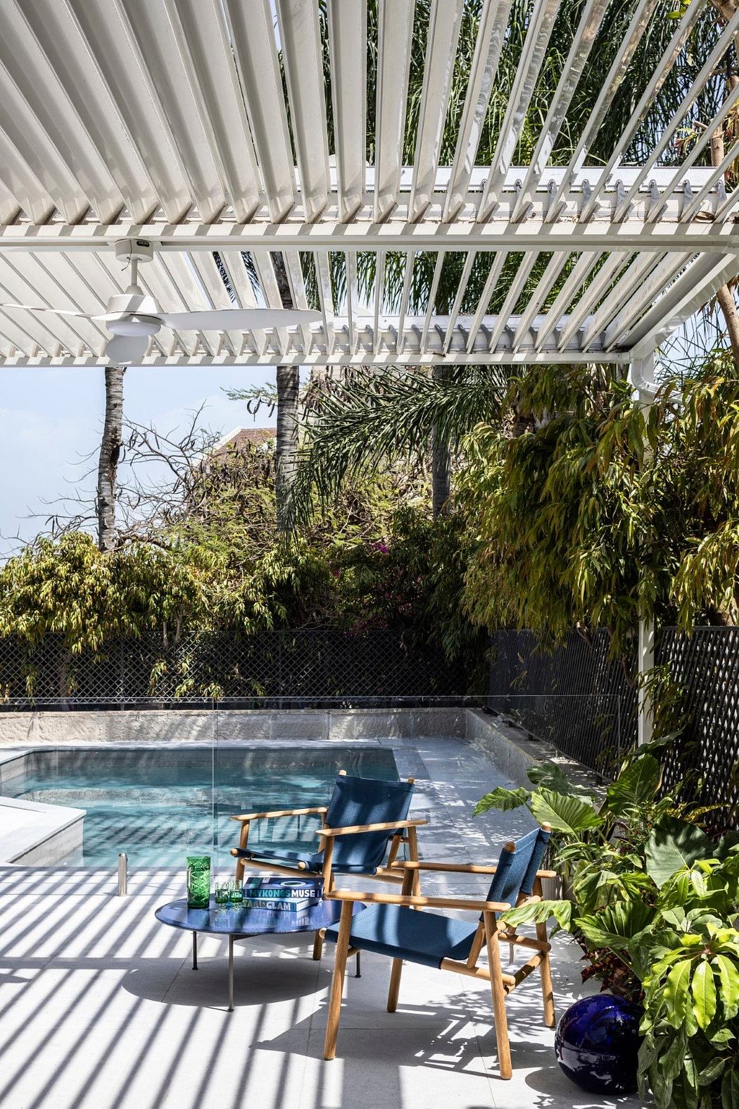 An outdoor patio with a pergola, pool, and modern patio furniture surrounded by lush greenery.