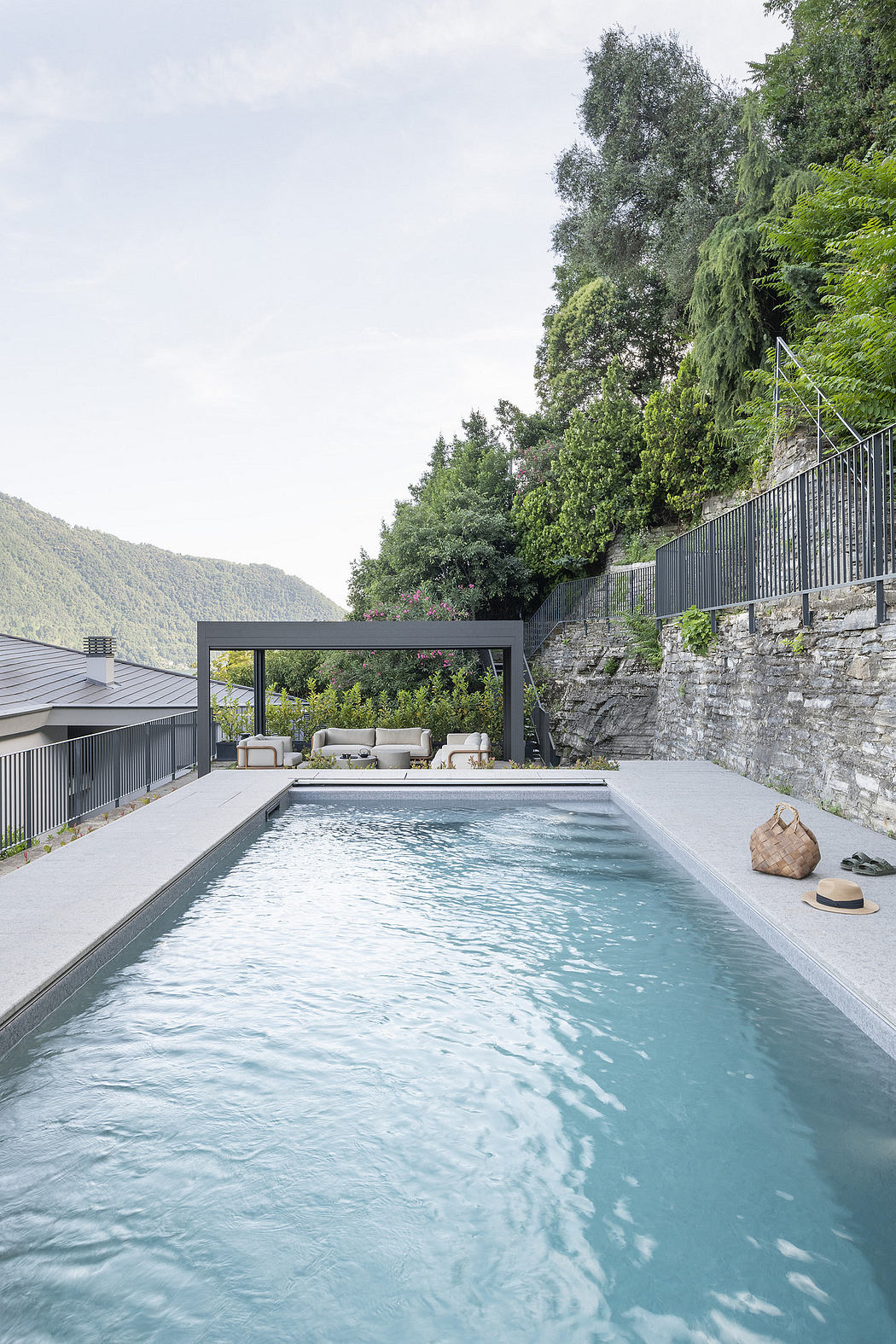 Sleek, modern pool area with stone wall, metal framing, and lush greenery backdrop.