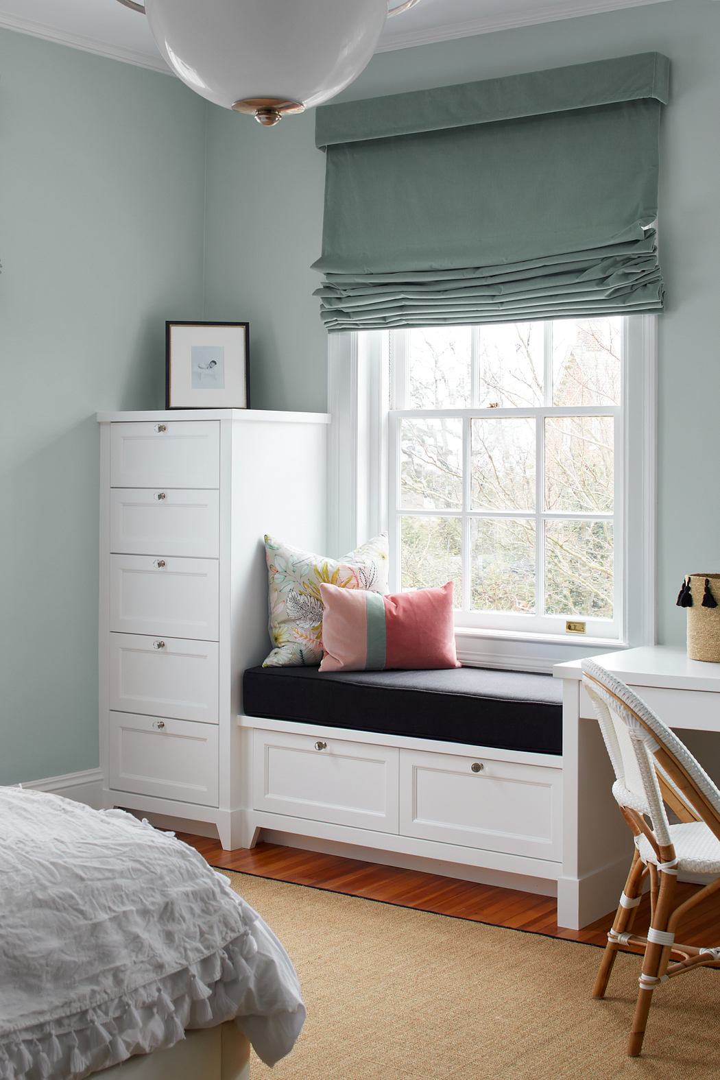 A cozy bedroom with a white storage cabinet, a window seat, and a gray Roman shade.