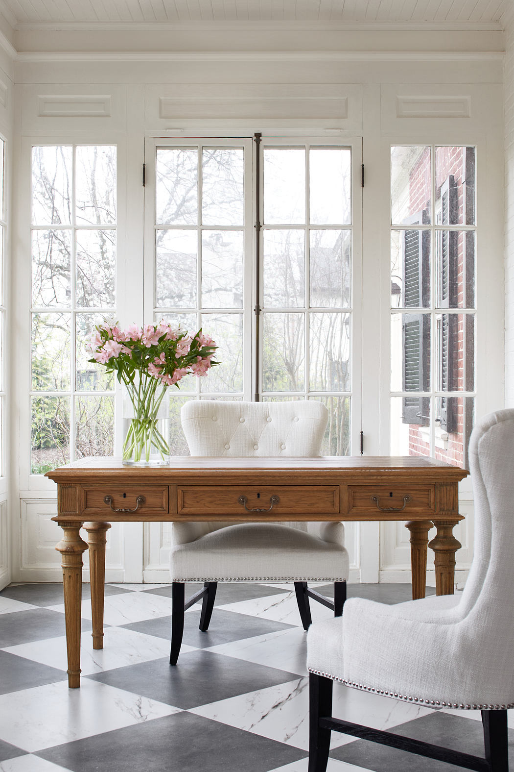 A bright, airy room with large windows, a wooden desk, and tufted white chairs on a black-and-white tiled floor.