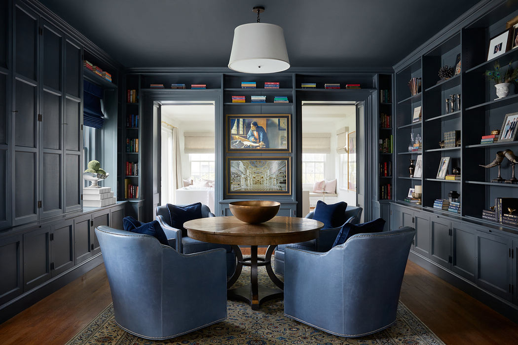 A cozy study with built-in bookshelves, a round wooden table, and plush blue chairs.