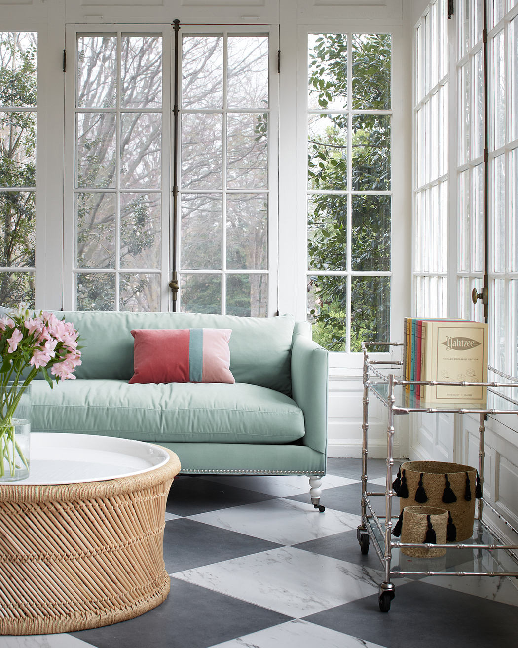 Bright, airy room with large windows, mint green sofa, pink accents, and a woven rattan coffee table.
