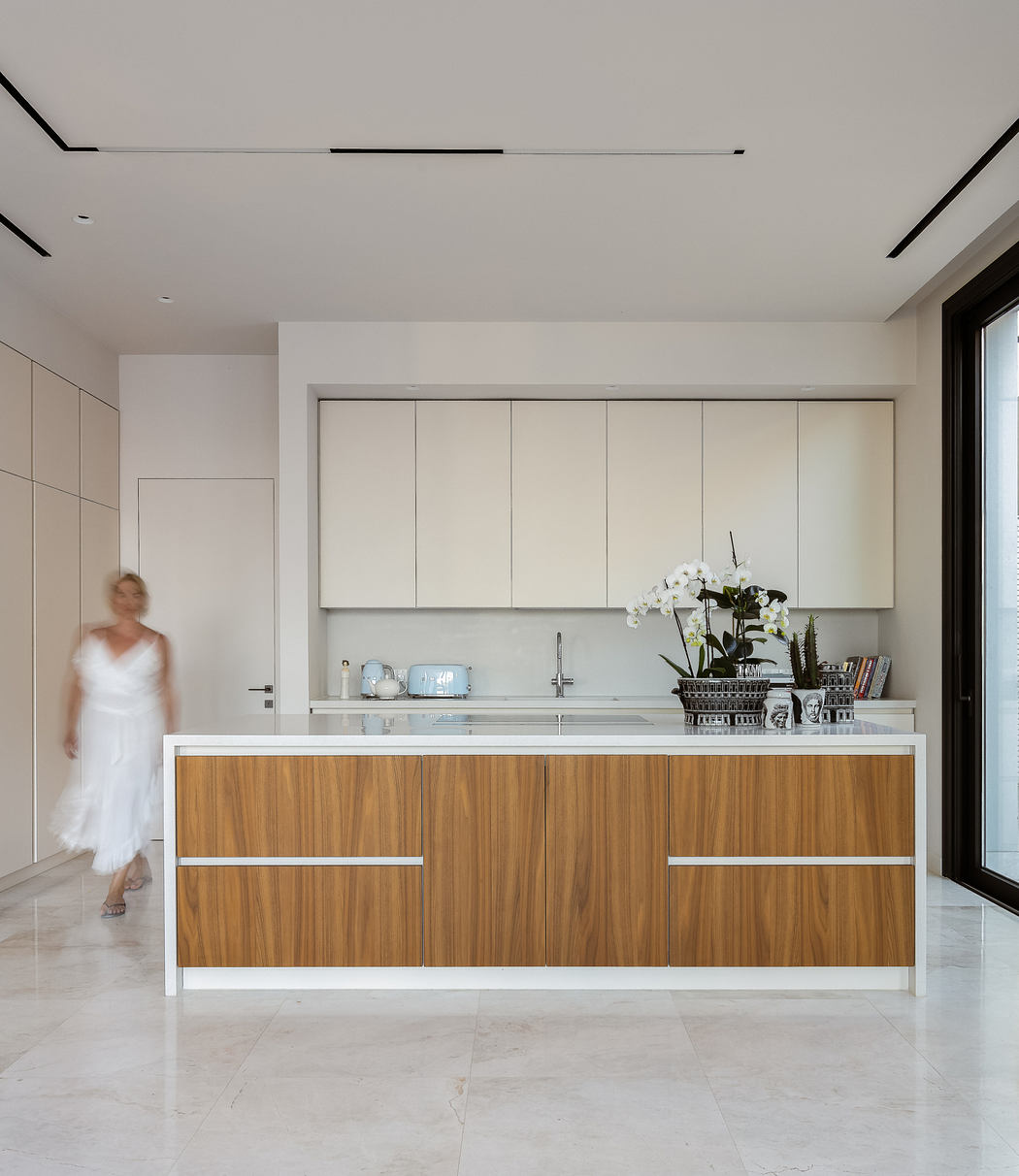 Minimalist kitchen with sleek white and wood cabinets, a central island, and a blurred person in the background.