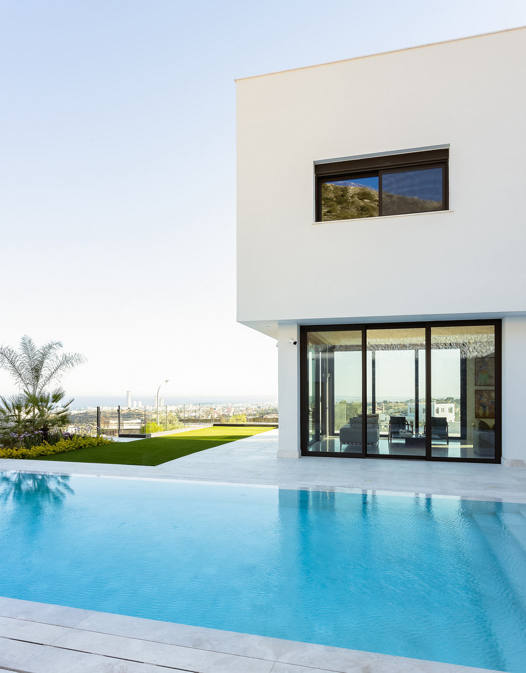 Modern minimalist architecture with a sleek pool, lush green lawn, and cityscape view.