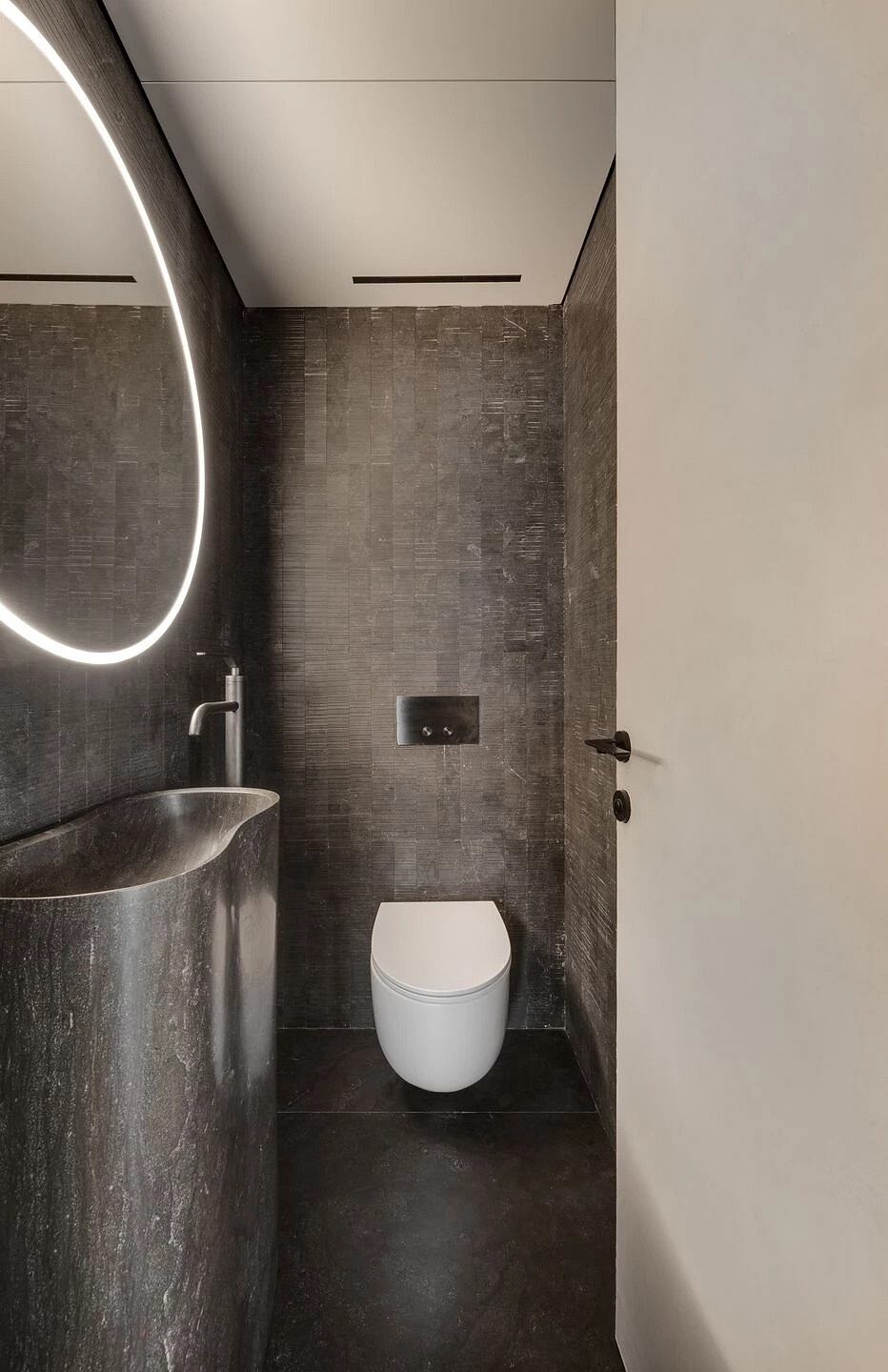 Modern bathroom with minimalist design: sleek toilet, oval mirror, and dark tile walls.