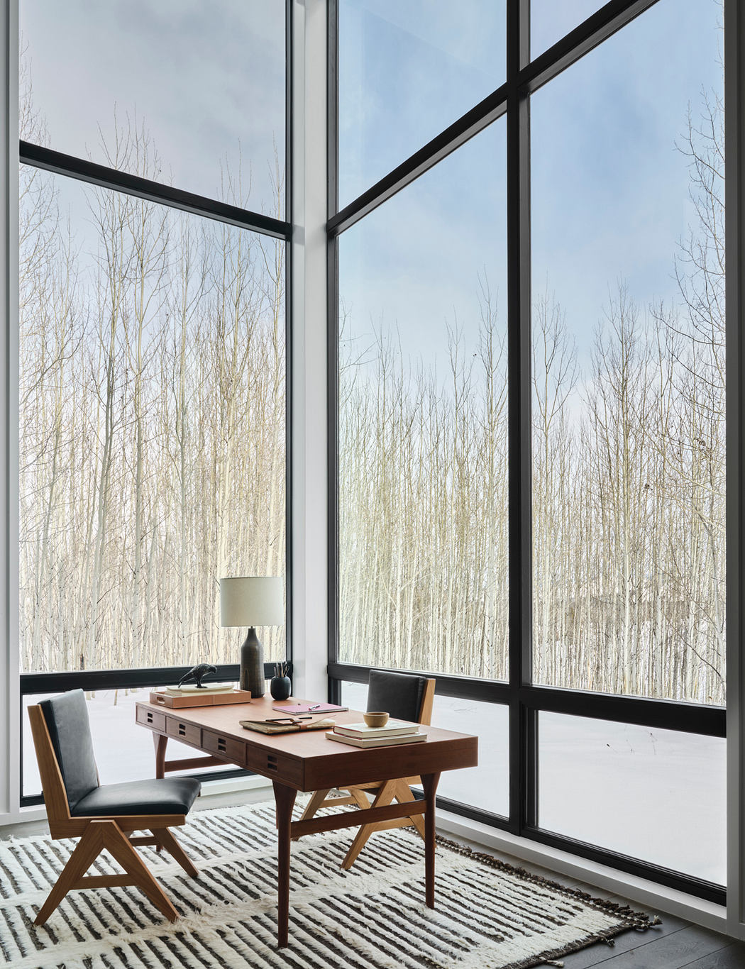 Minimalist workspace with floor-to-ceiling windows framing a wintry landscape, accented by a wooden desk and chair.