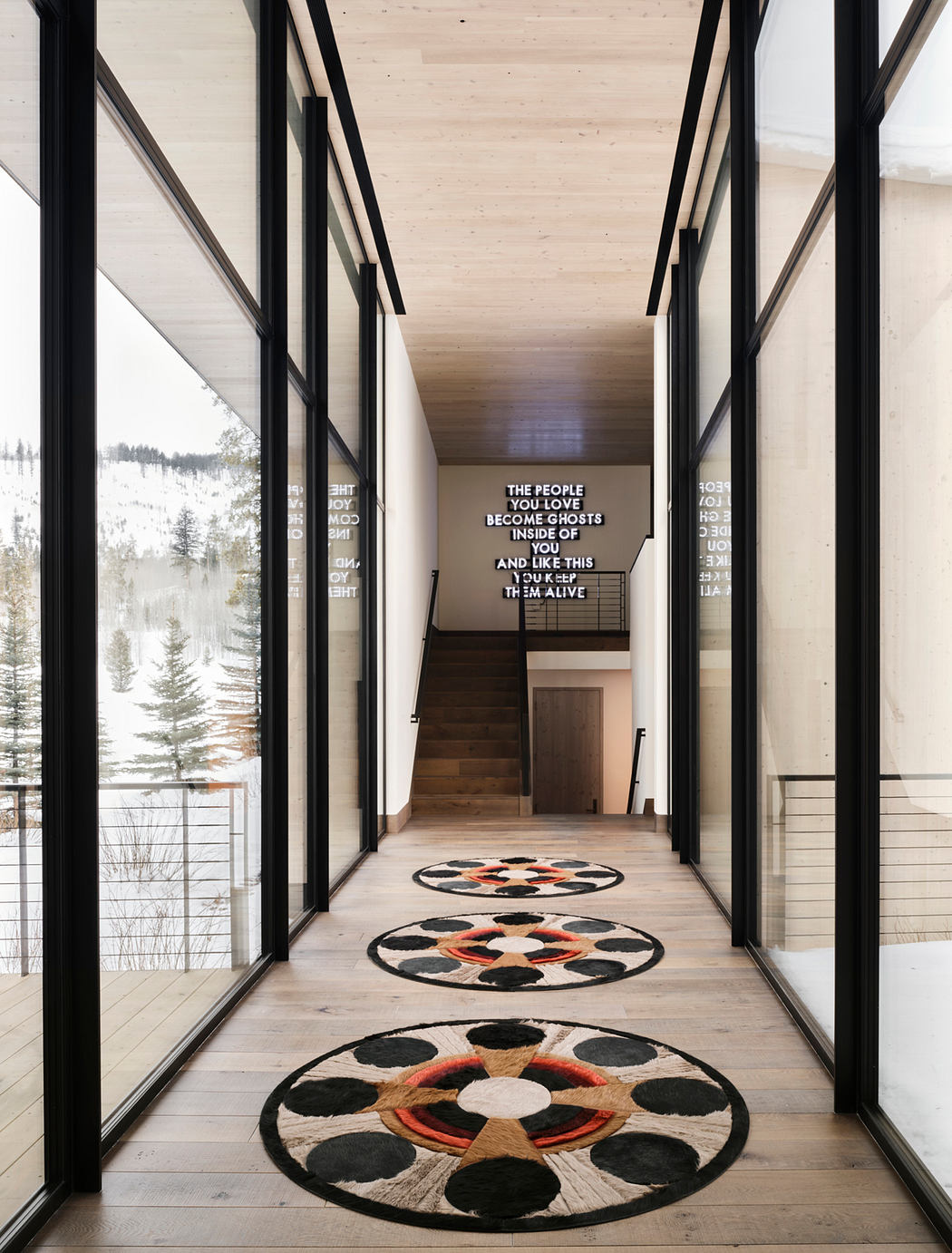 A modern, open-concept hallway with large windows, wood floors, and patterned rugs leading up to stairs.