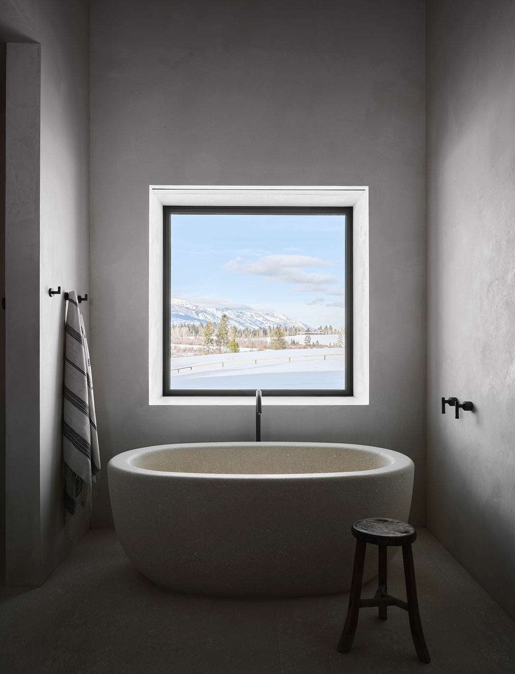 A modern, minimalist bathroom with a freestanding tub overlooking a snowy mountain landscape through a large window.