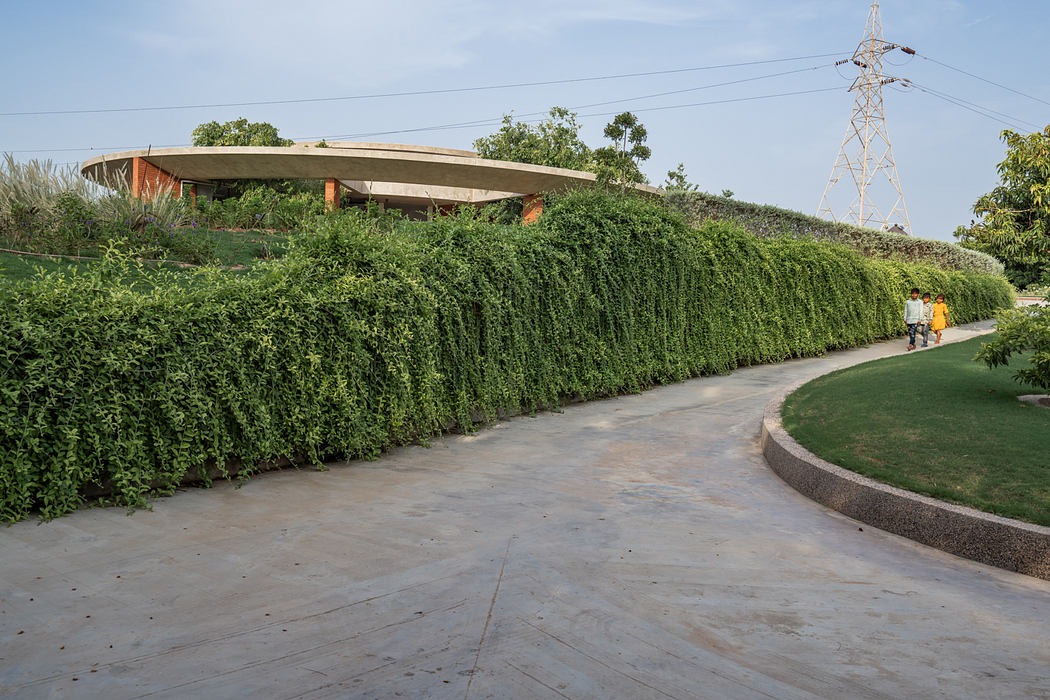 The image depicts a modern, curved concrete structure surrounded by lush greenery and paved walkways.