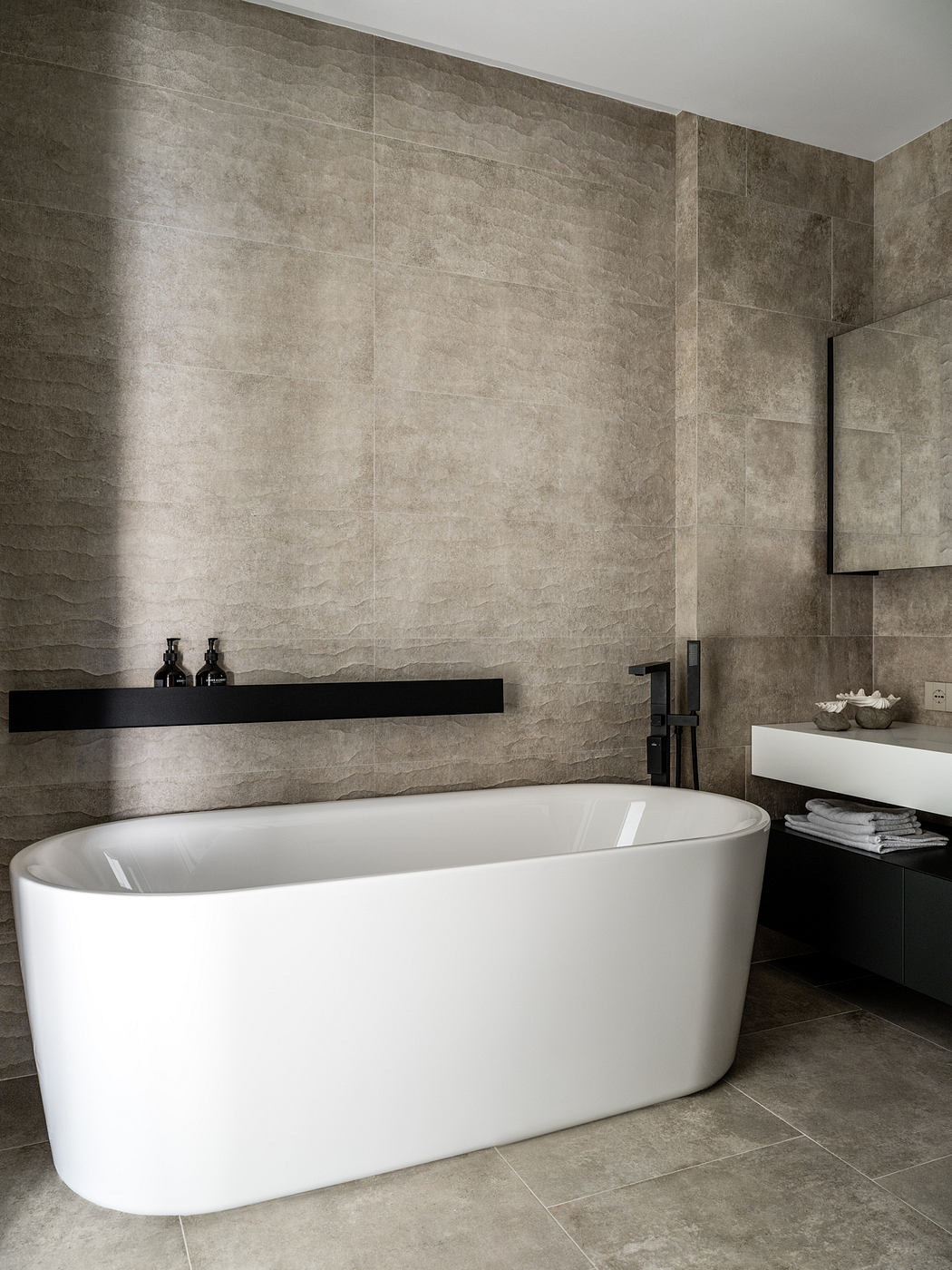 Minimalist bathroom design with white freestanding tub, dark tile walls, and shelving.