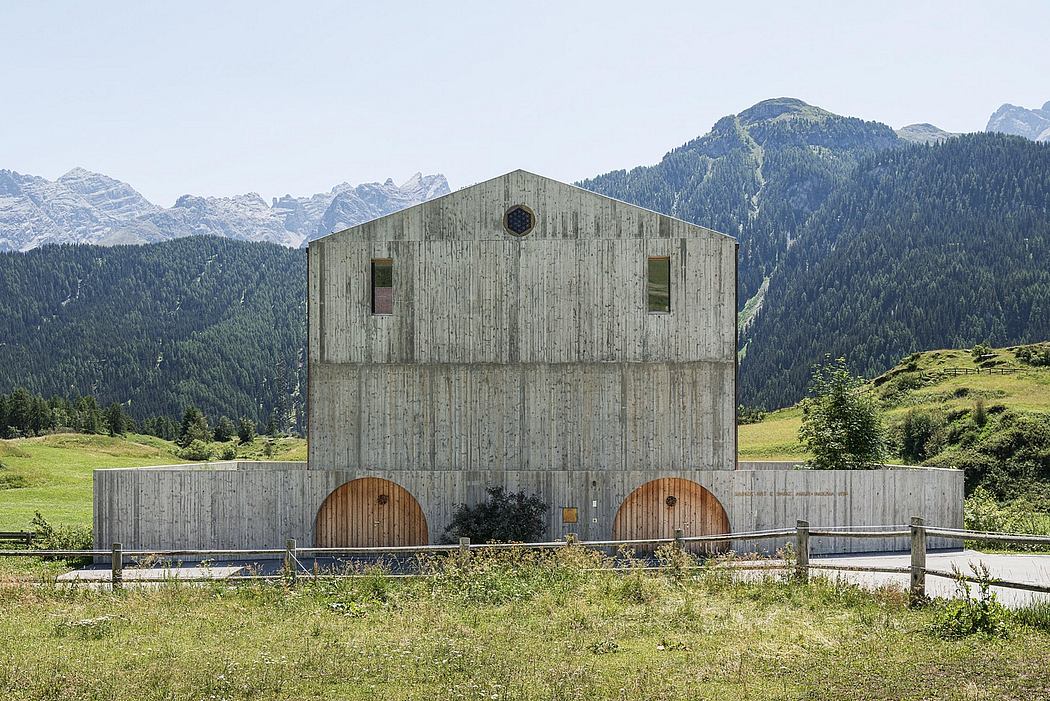 A large, gray wooden barn-like structure with two circular windows and two circular entrances against a mountainous backdrop.