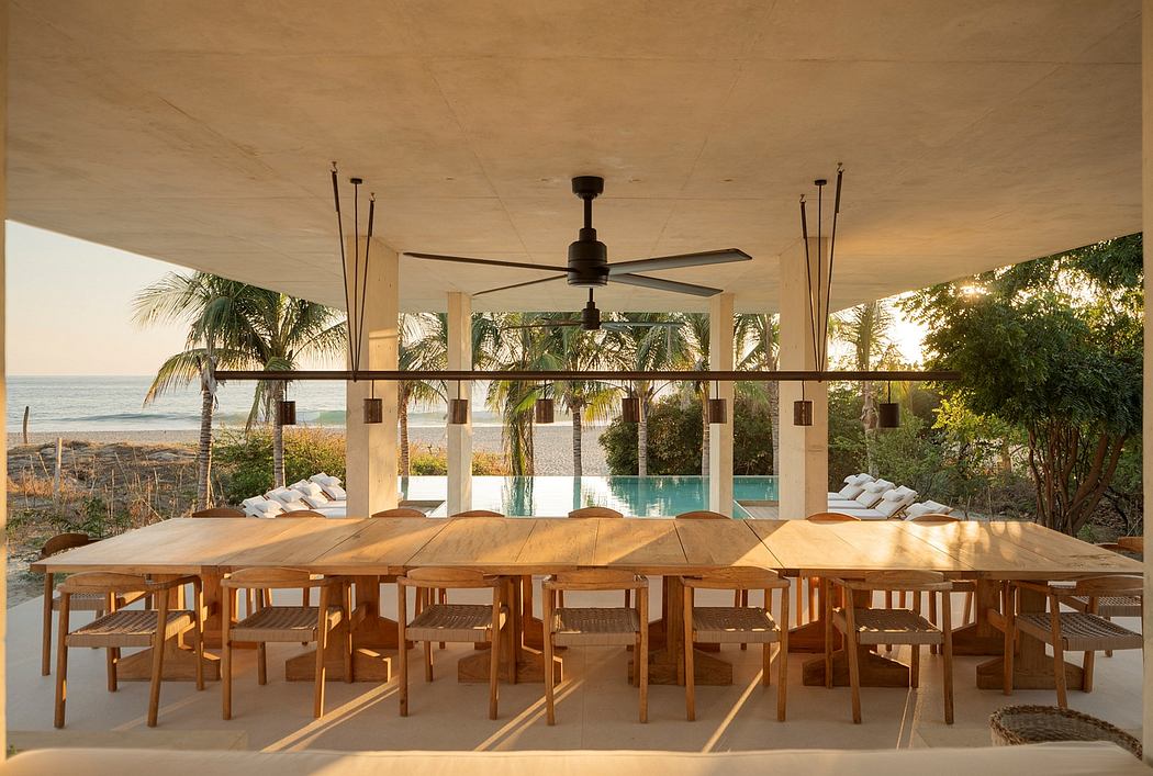 A covered outdoor dining area with a large wooden table, ceiling fan, and views of a pool and palm trees.
