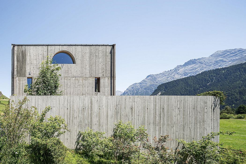 The image shows a modern, concrete-and-wood building nestled in a lush, mountainous landscape.