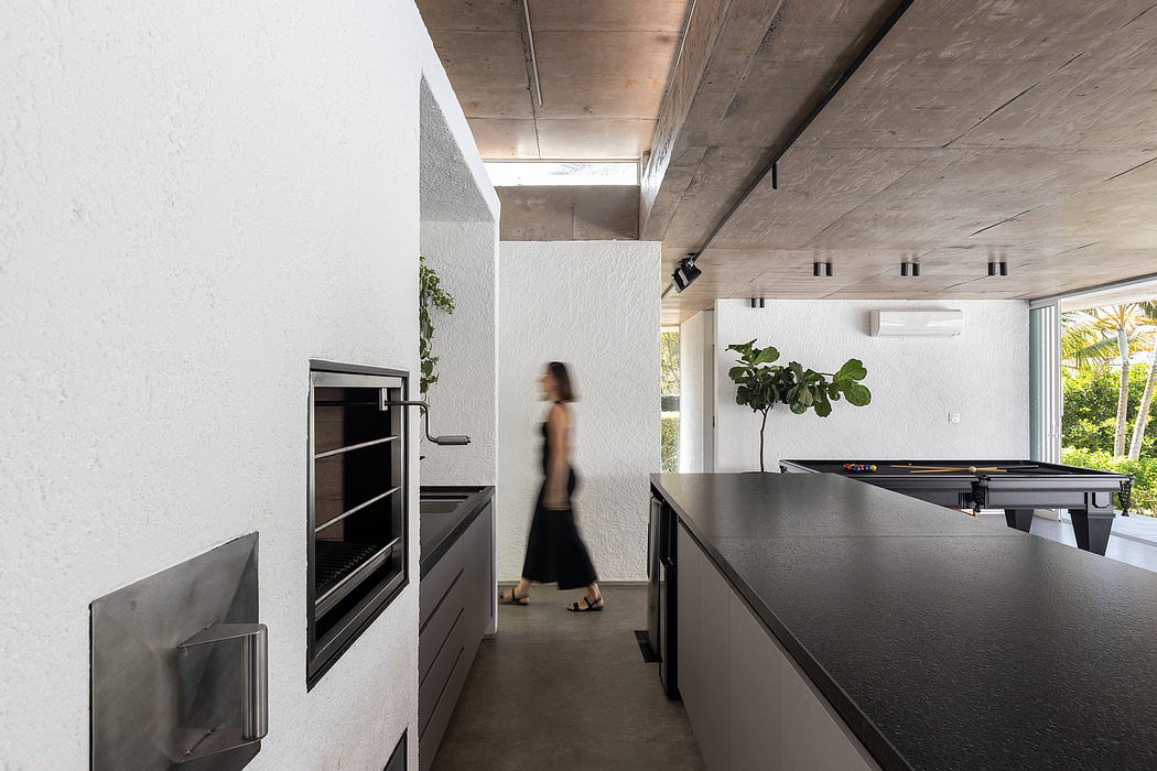 A modern, minimalist kitchen with sleek black cabinetry, concrete ceiling, and a person walking through.