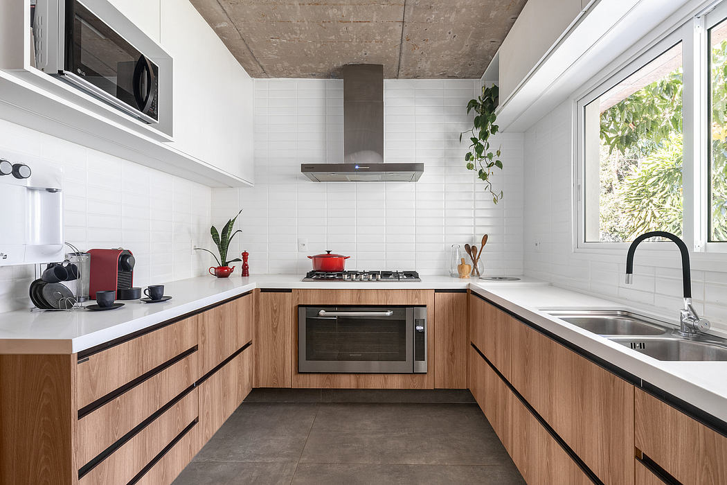 A modern kitchen with white tiles, wood cabinets, and concrete ceiling. Black appliances and a sink with large windows.