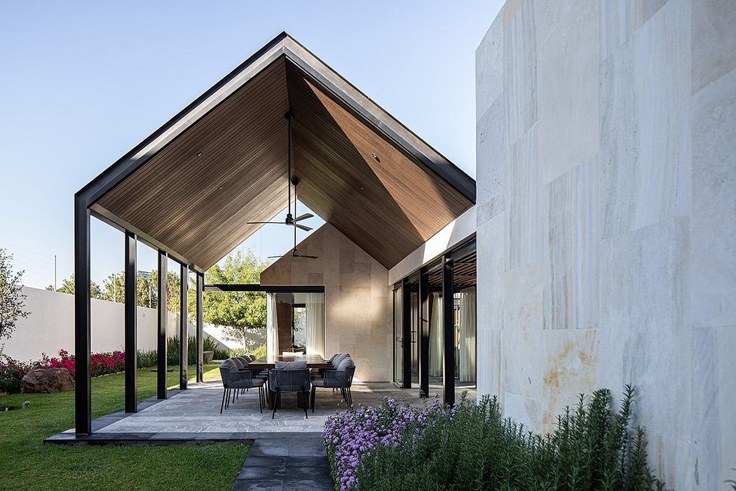 A modern open-plan patio with a wooden-beamed roof and large sliding glass doors, surrounded by a lush garden.