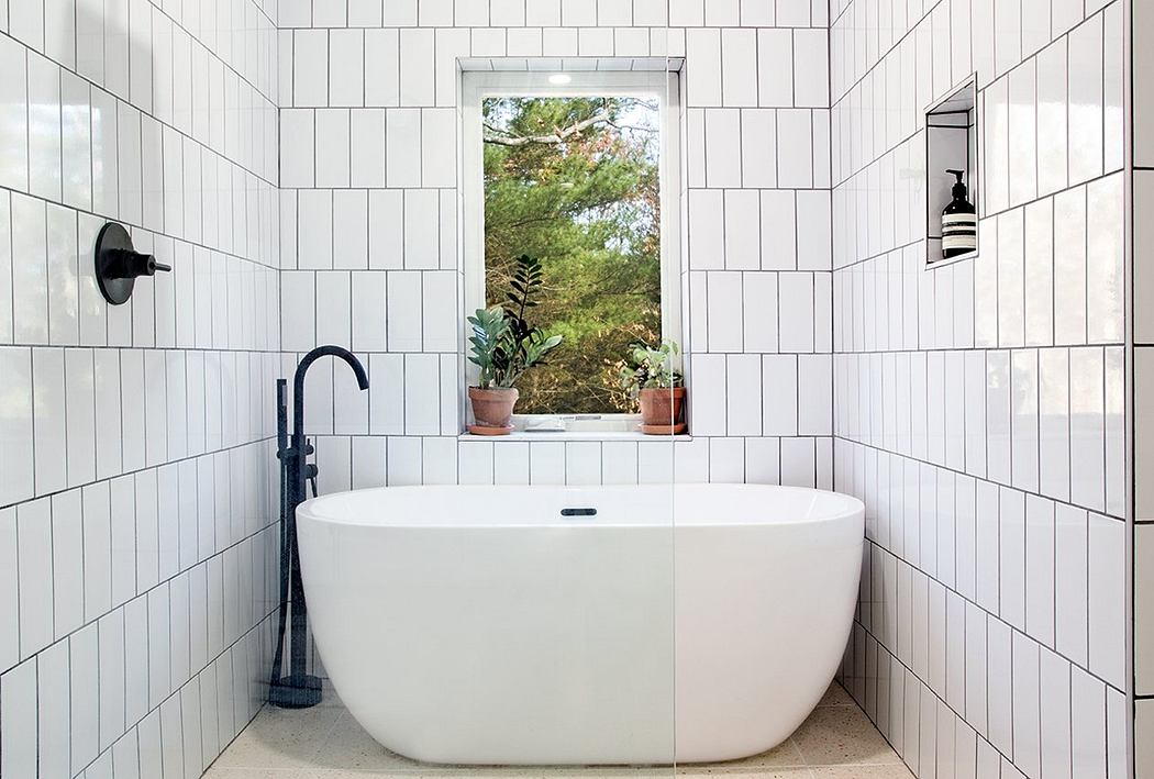 A modern bathroom with a freestanding tub, square white tiles, and a window overlooking nature.