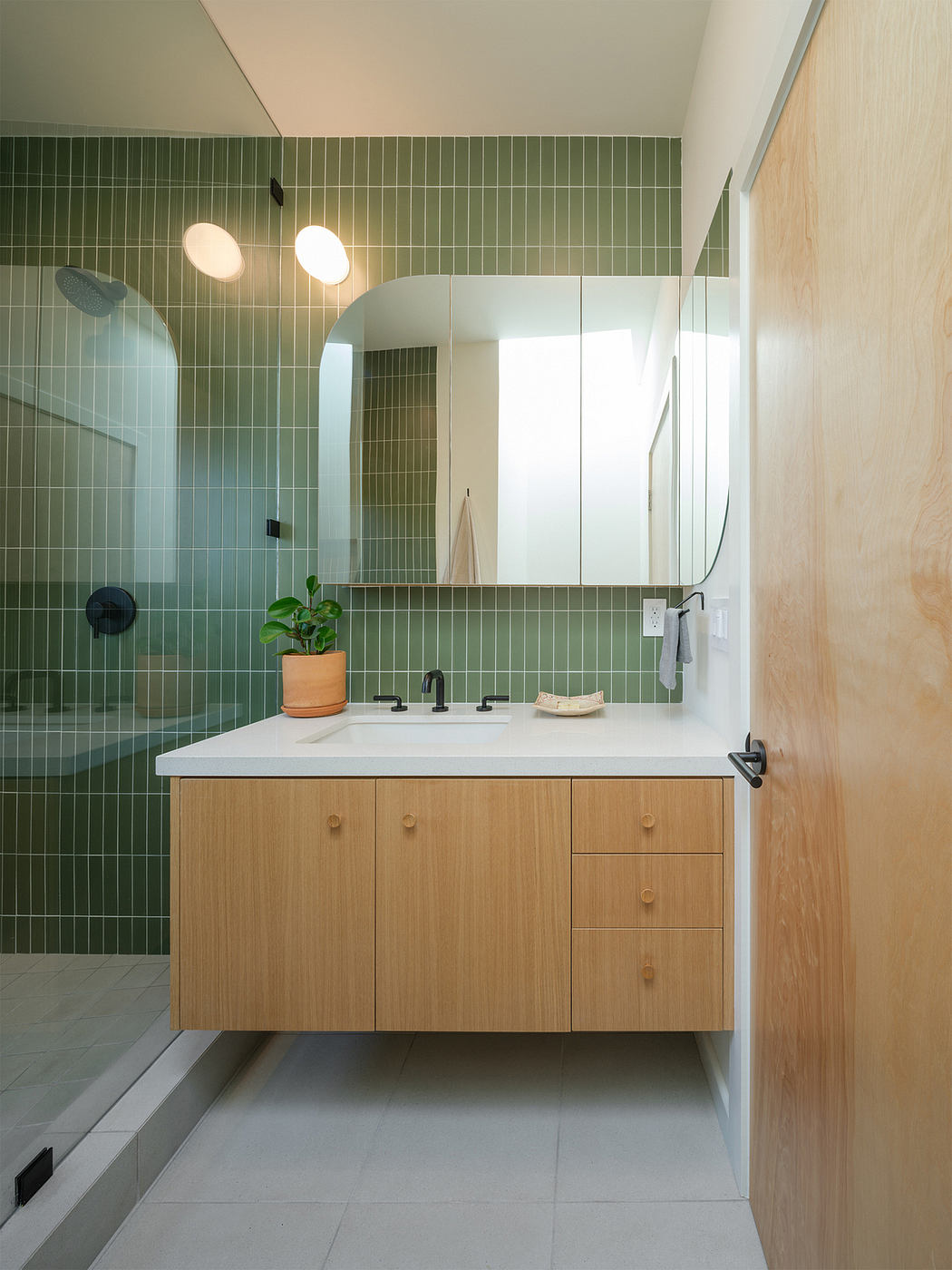 A contemporary bathroom with a wooden vanity, green tile walls, and recessed lighting.